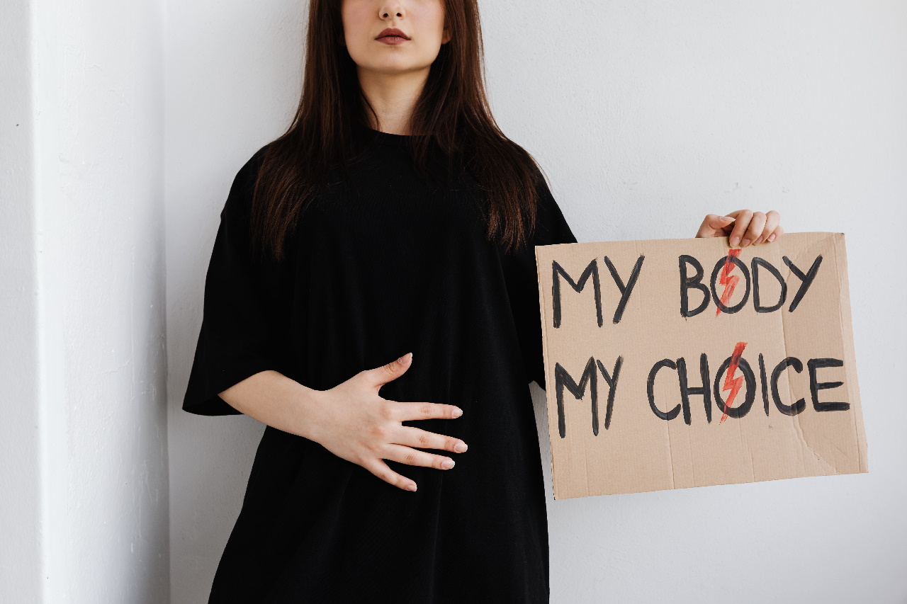 Frau in schwarzem Kleid, die sich den Bauch hält und daneben ein Schild mit der Aufschrift "My body, my choice" hält