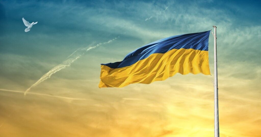 Fahne der Ukraine weht vor einem Himmel im Hintergrund in ukrainischen Nationalfarben, links oben ist eine Friedenstaube zu sehen