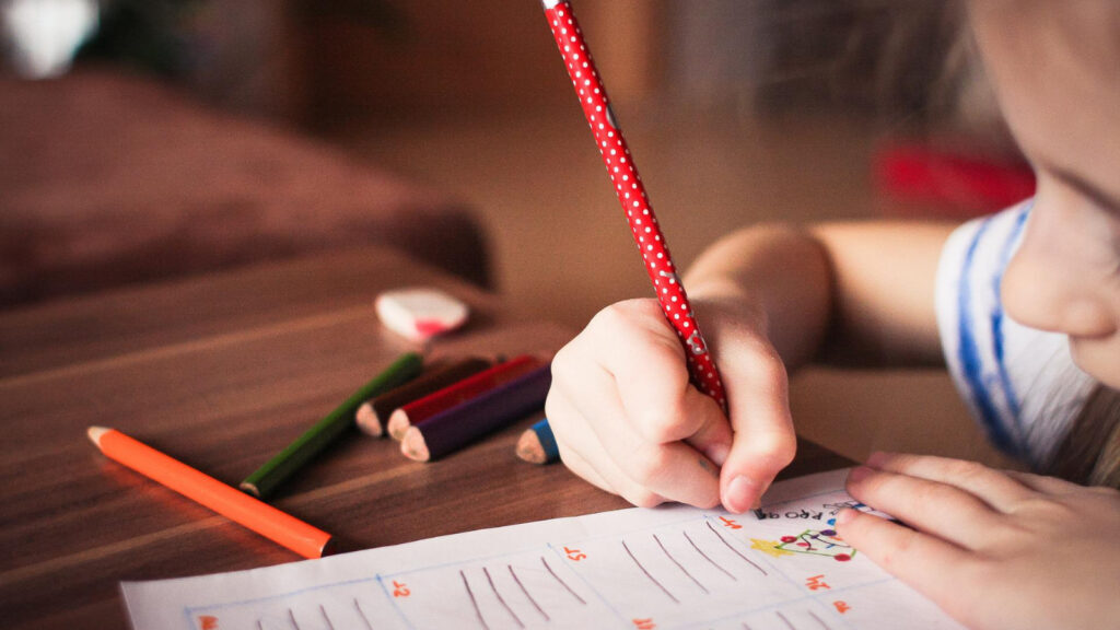 Schulkind malt mit großem Bleistift auf einem Aufgabenheft (Nahaufnahme)