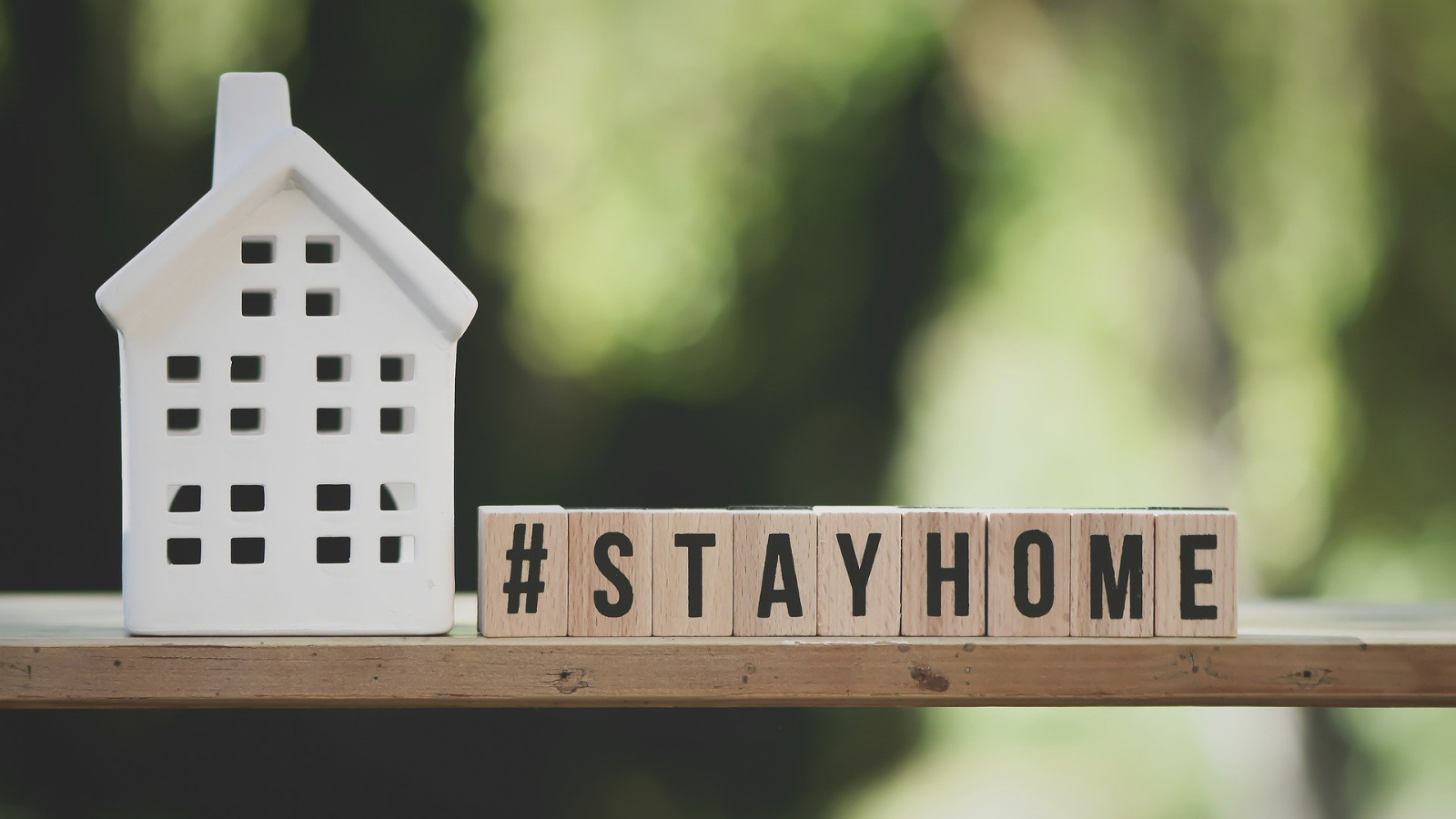 Symbolbild: auf einem Regalbrett steht vor einem grünlichen Hintergrund links ein stilisiertes Haus aus Ton oder Plastik und rechts ist mit beschrifteten Holzklötzen der Hashtag #STAYHOME abgebildet